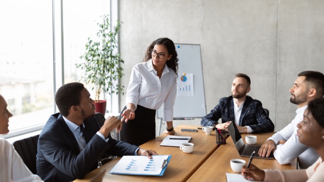 A standing woman leading a group of people in a meeting.