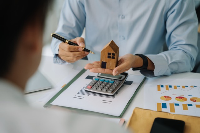 Close up of someone sitting at a desk covered in charts and graphs. They are holding a small wooden house in one hand and gesturing to it with the other.