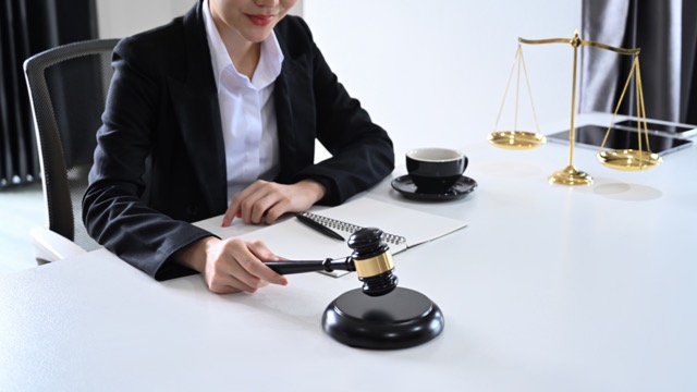 A person sitting at a desk hitting a gavel on a block, a pair of scales in the background, and an open notebook with a pen on the desk.