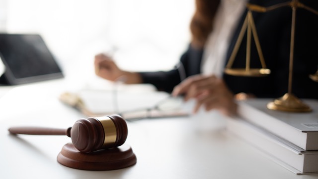 A gavel resting on a block with a pair of scales and a person with open notebook in the background.