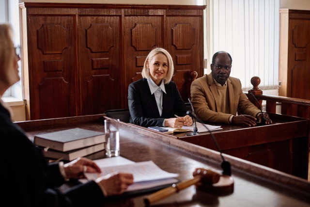 An attorney sitting next to a client in a courtroom, speaking to a judge.